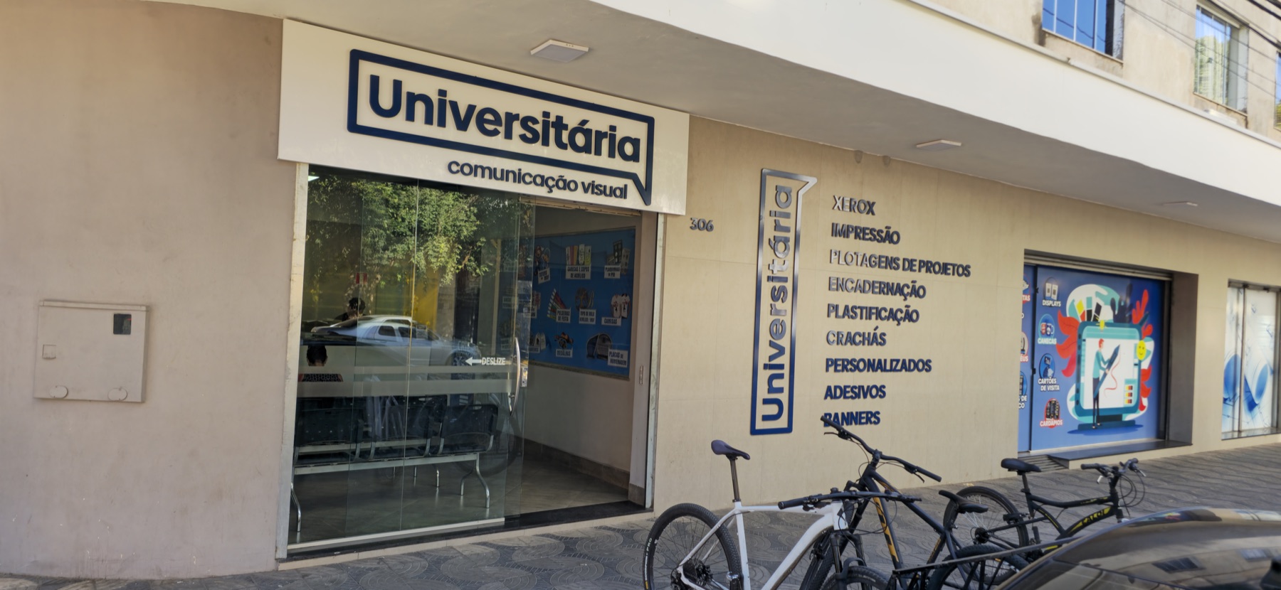 Fachada da Universitária Comunicação Visual no número 306 da Rua José Luiz Nogueira, no Centro de Governador Valadares, com o letreiro da loja e a lista de serviços aplicada na parede