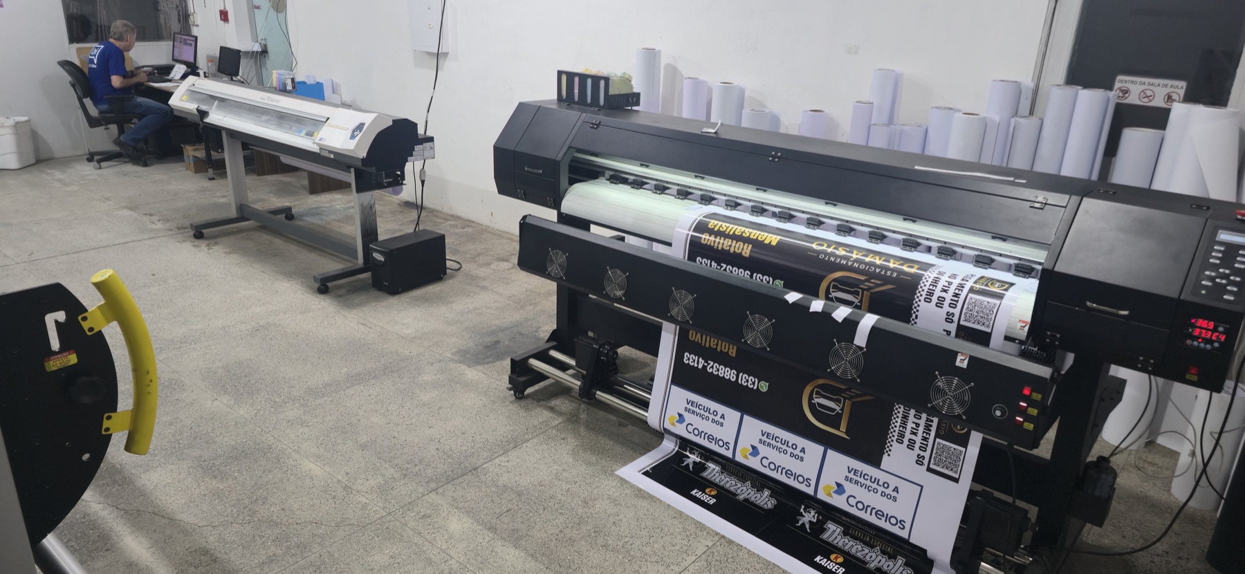 Impressora de grande formato da Universitária com adesivos impressos saindo da máquina, ao lado dos rolos de material e da mesa de arte-final