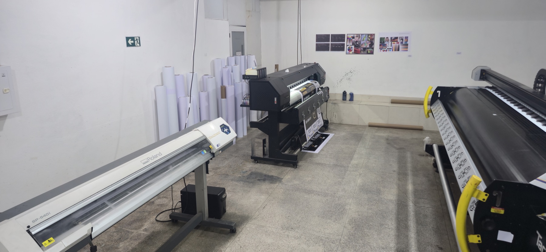 Sala de produção da Universitária com plotter de recorte Roland, impressora de grande formato e rolos de material encostados na parede
